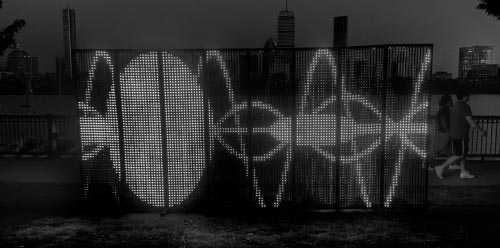 large outdoor screen on a river bank, with people walking by, showing abstract graphics