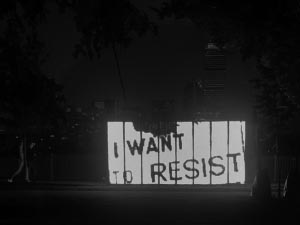 large outdoor screen on a river bank, with a person walking by, showing text that reads: I want to resist